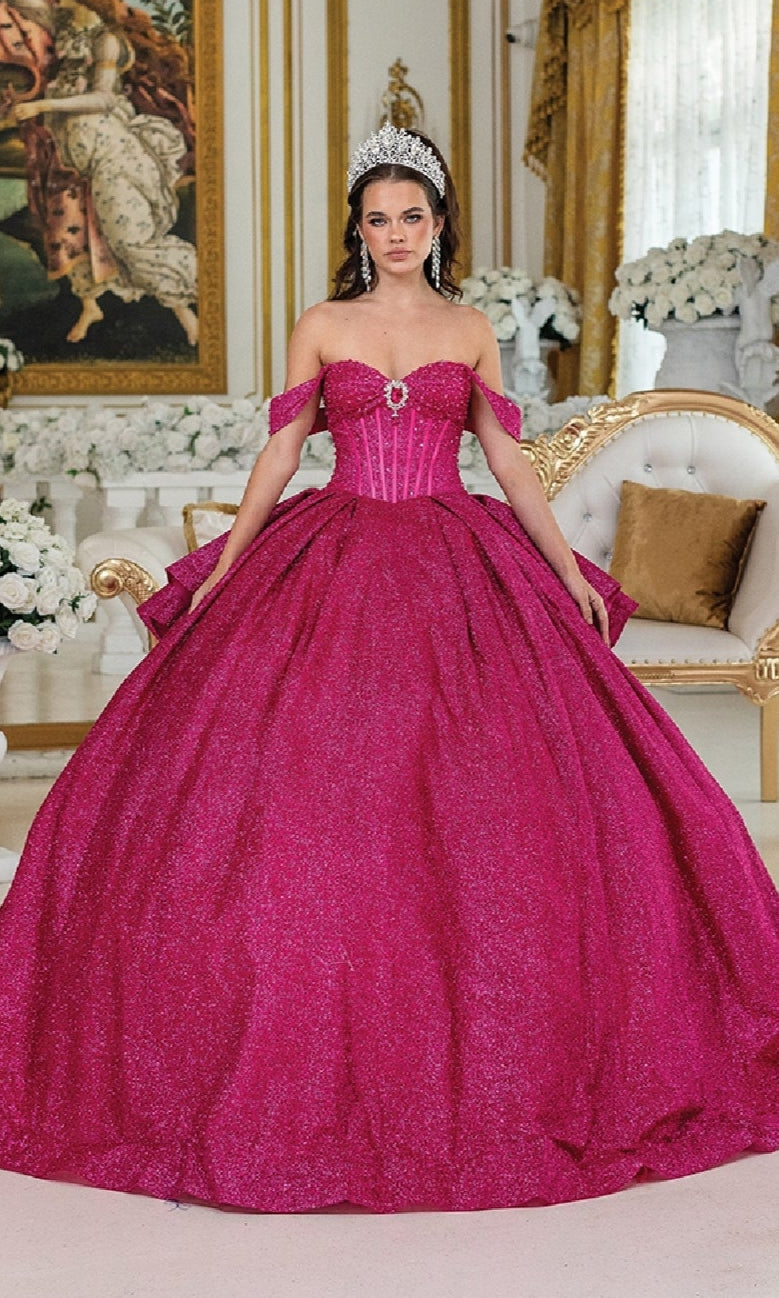 A woman in the Quinceanera Dress 1937 By Dancing Queen, a sparkling bright pink ball gown with off-the-shoulder straps, wears a silver tiara and stands in an ornate room decorated with white flowers and gold accents.